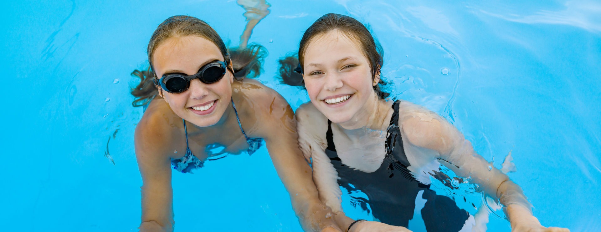 Two girls smiling in a swimming pool, one wearing goggles, enjoying a sunny day.