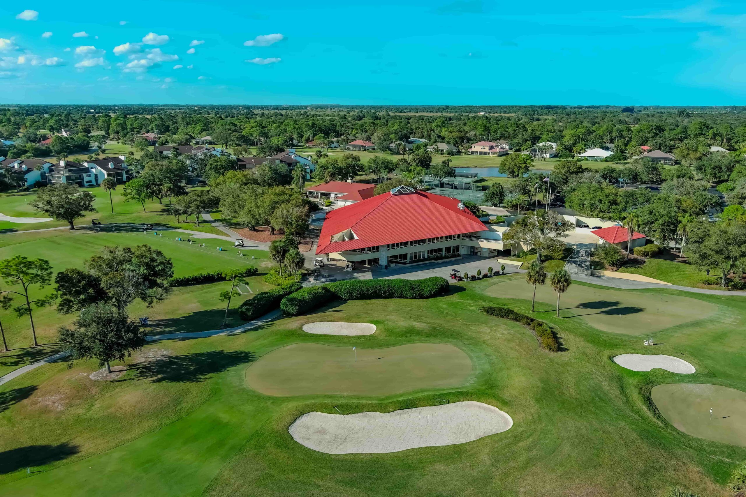 Aerial view of a golf clubhouse with a red roof surrounded by green lawns and trees, with golf carts in the distance.