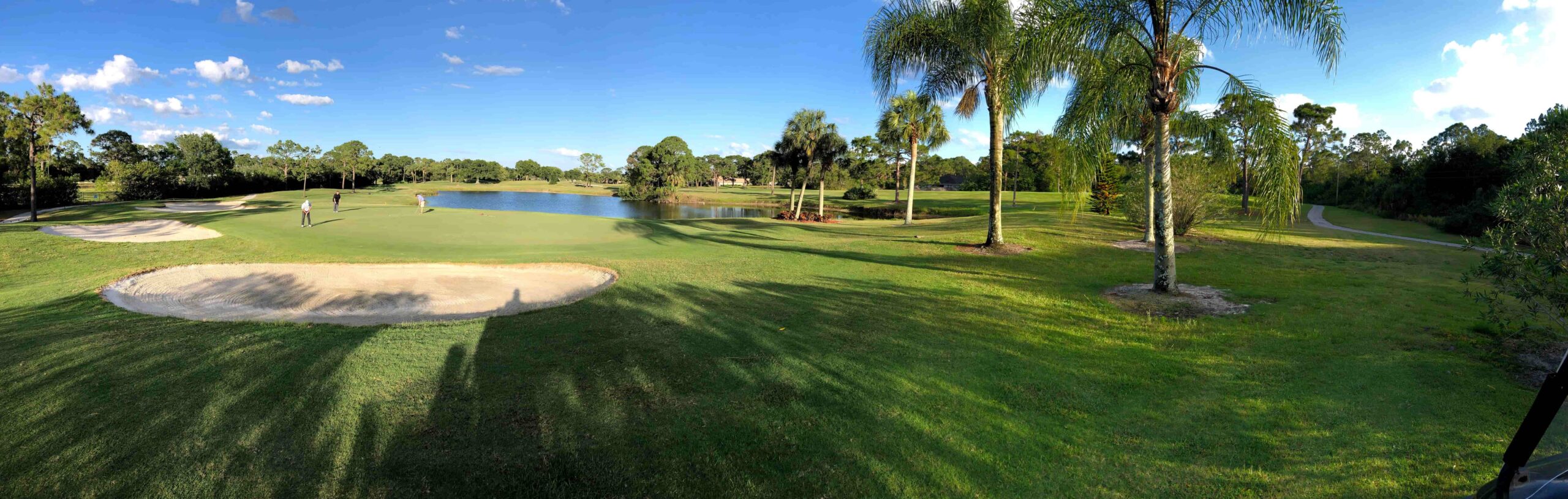 Golf course scene with two players on the green, sand traps, and a pond under a blue sky with clouds.